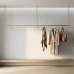 Tringle de penderie minimaliste en laiton doré fixée au plafond au-dessus d'un banc en bois dans une entrée épurée.