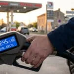 Une main tenant un pistolet de pompe à essence avec un écran numérique affichant un prix élevé de 2,45 € par litre.