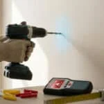 Mains avec gants perçant un mur en placo avec une perceuse sans fil, près d'un détecteur de matériaux et de chevilles.
