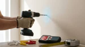 Mains avec gants perçant un mur en placo avec une perceuse sans fil, près d'un détecteur de matériaux et de chevilles.