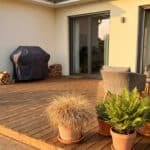 Terrasse en bois avec barbecue couvert, plantes en pot et salon de jardin, au soleil couchant.