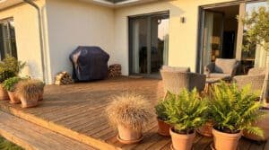 Terrasse en bois avec barbecue couvert, plantes en pot et salon de jardin, au soleil couchant.
