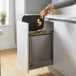 Main jetant des épluchures dans un bac à compost en inox coulissant intégré sous un plan de travail de cuisine moderne.
