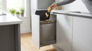 Main jetant des épluchures dans un bac à compost en inox coulissant intégré sous un plan de travail de cuisine moderne.